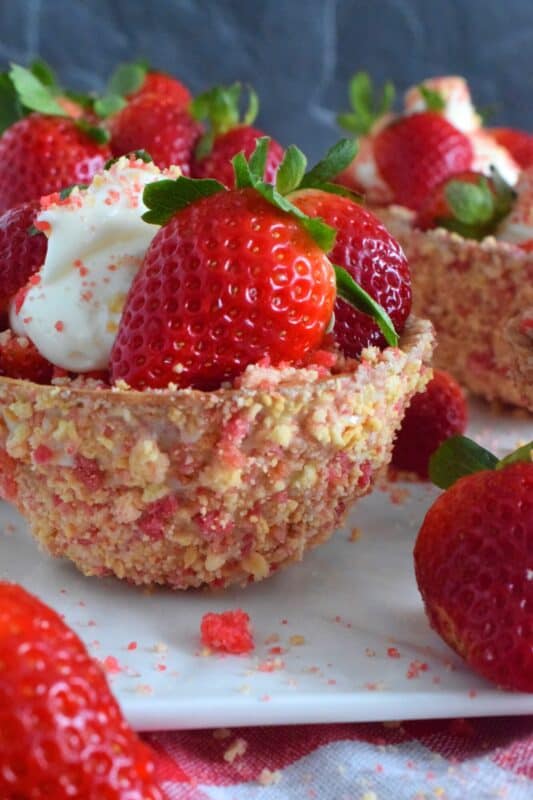 Sugary sweet and crunchy waffle bowls are coated with strawberry shortcake crumbs and filled with a light and fluffy cheesecake mixture, topped with fresh strawberries, and more strawberry shortcake crumbs. This gorgeously impressive dessert is for the serious strawberry lover! #shortcake #waffle #strawberry #cheesecake
