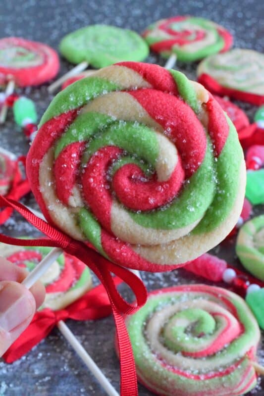 Add colour and pizzazz to your holiday cookie platter with these Holiday Lollipop Cookies! Spiralled and rolled cookie dough is topped with sparkling sugar and baked with a stick. These are sure to produce lots of smiles and crown you the winner of holiday baking! #lollipop #cookies #christmas