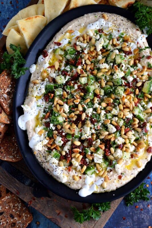 What's a party without dip? Hummus Party Dip Platter is a large, layered dip that's just perfect for entertaining a crowd. This all-in-one dip has hummus, sour cream, pine nuts, sun dried tomatoes, cucumbers, feta, and a few other ingredients too! Serve this on any occasion for a delicious, crowd-pleasing appetizer! #hummus #dip #appetizer #party #platter #spread